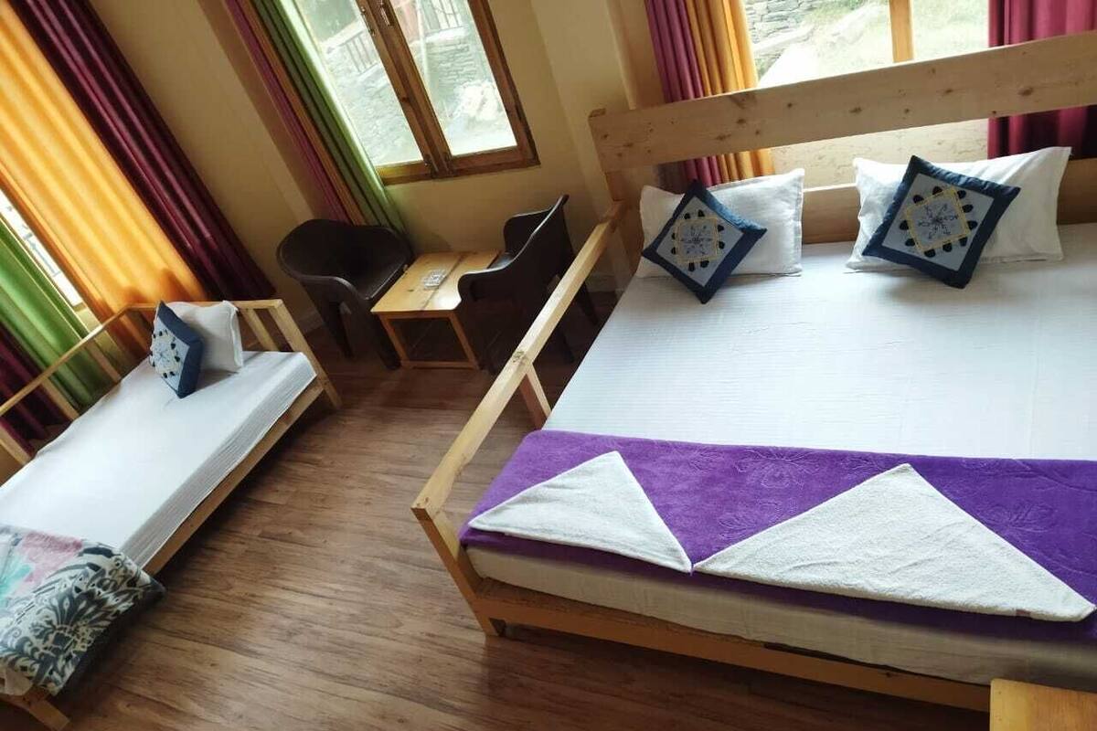 Ein hölzernes Bett mit einer violett-weißen Tagesdecke, zwei Kissen mit Uhrendruck, eine Holzbank, ein kleiner Tisch und ein Stuhl.