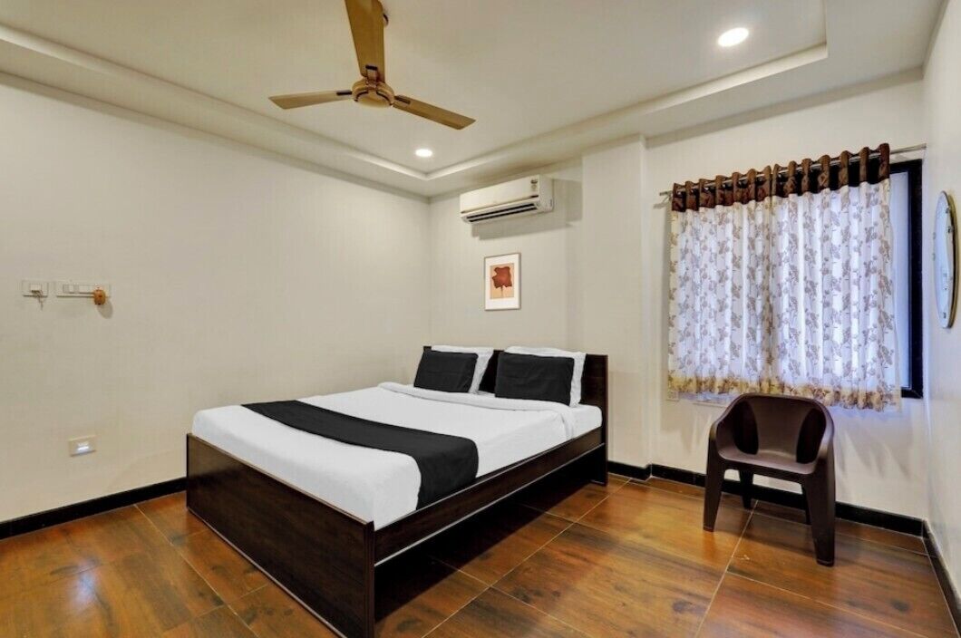 A bedroom with a bed, a ceiling fan, an air conditioning unit, a window with curtains, and a small stool.