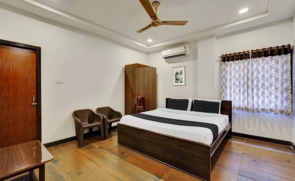 A bedroom with a wooden bed, a ceiling fan, an air conditioning unit, a wardrobe, two plastic chairs, and a window with floral curtains. - MEDI KONDA GRAND (Nellore)