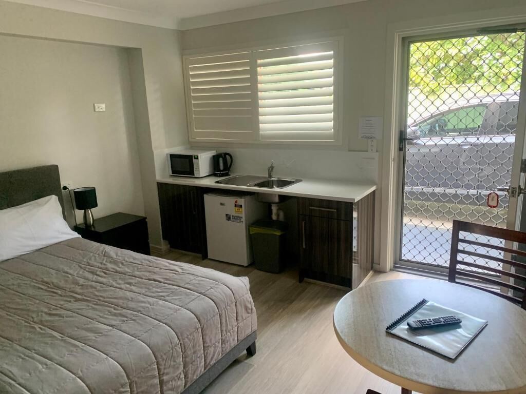 Un dormitorio con cama, una kitchenette pequeña, una mesa de comedor con un control remoto y vistas a través de una ventana con persianas.