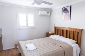Une chambre à coucher comprenant un lit, une table de chevet, un climatiseur fixé au mur, un ventilateur de plafond, une fenêtre et un tableau encadré accroché au mur.