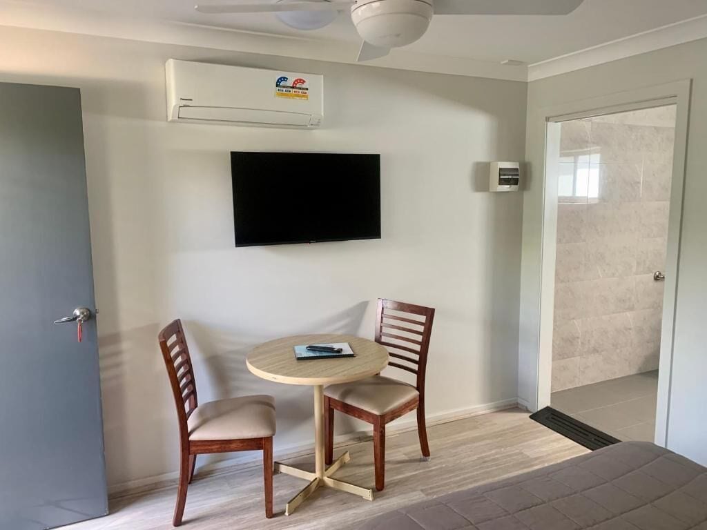 Quarto com televisão de tela plana na parede, uma mesa redonda com duas cadeiras e um ventilador de teto.