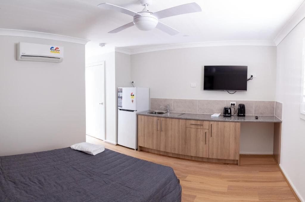 A compact room with a bed, kitchenette, ceiling fan, TV, and air conditioning unit.