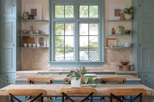 A kitchen with a large window, wooden chairs, and shelves with various items.