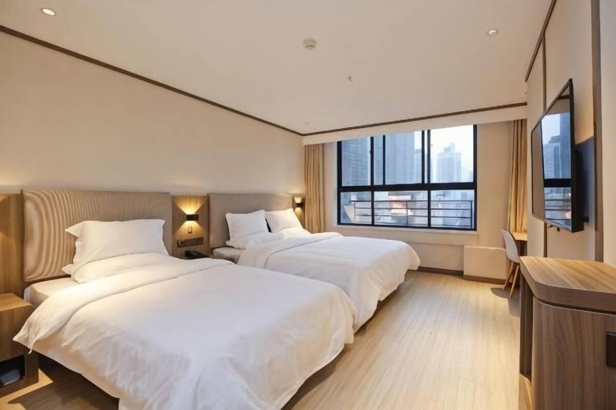 A hotel room with two beds, a TV, a window with a city view, and a desk.