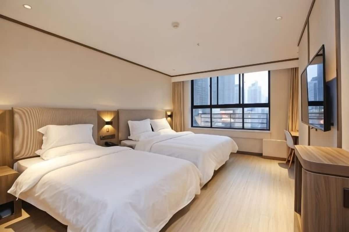 A hotel room with two beds, a TV, a window with a city view, and a desk.