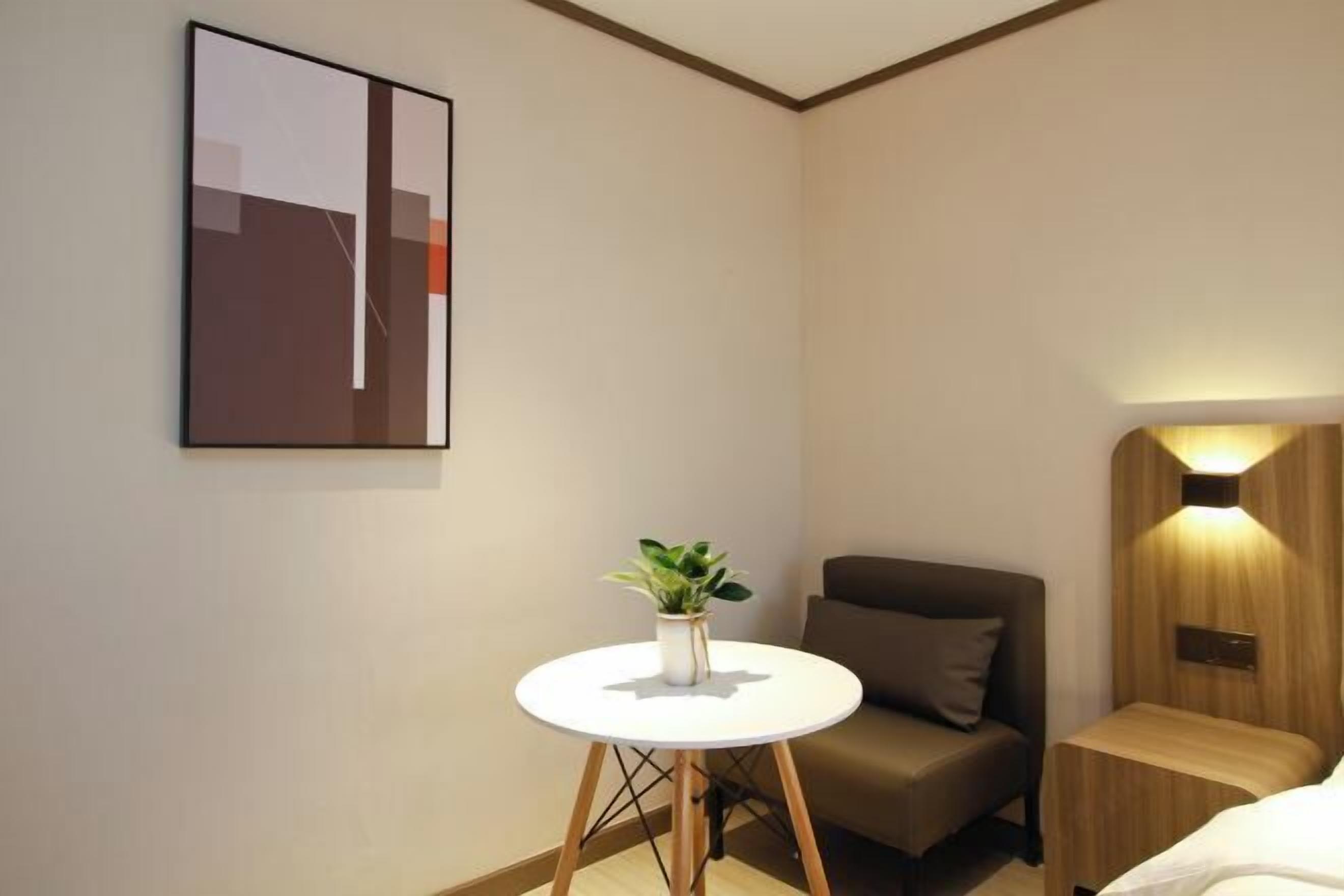 A modern hotel room with a single bed, a small round table with a plant, a brown armchair, and a wall-mounted light fixture.
