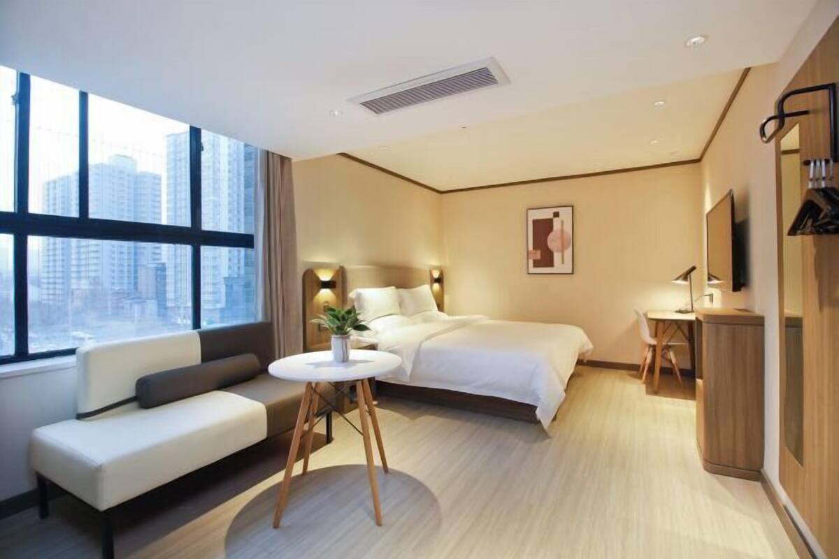 A modern hotel room with a bed, a sofa, a desk, and a large window with a city view.
