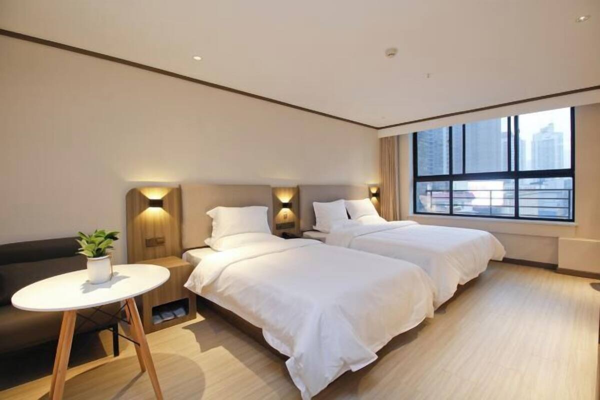 A hotel room with two beds, a small round table with a plant, and a view of buildings through large windows.