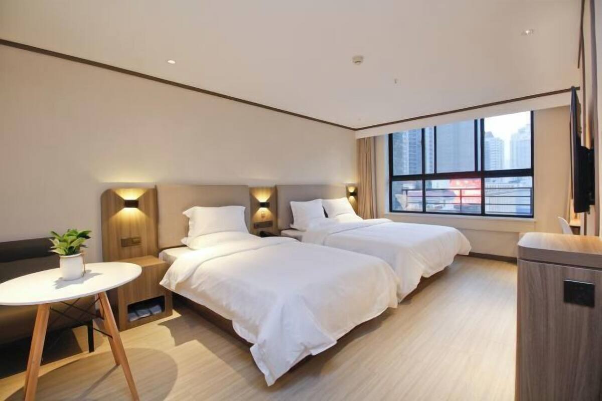 A hotel room with two beds, a small table, a plant, and a window with a city view.