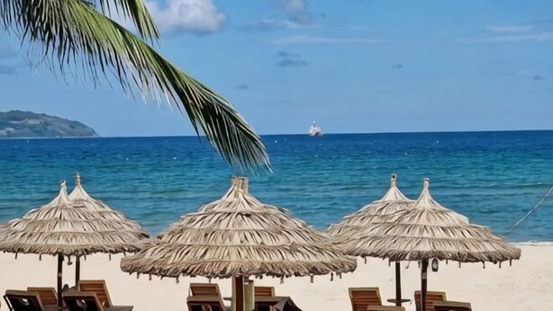 Une plage avec des parasols en chaume et des chaises longues, une mer d’un bleu limpide et un navire à l’horizon.