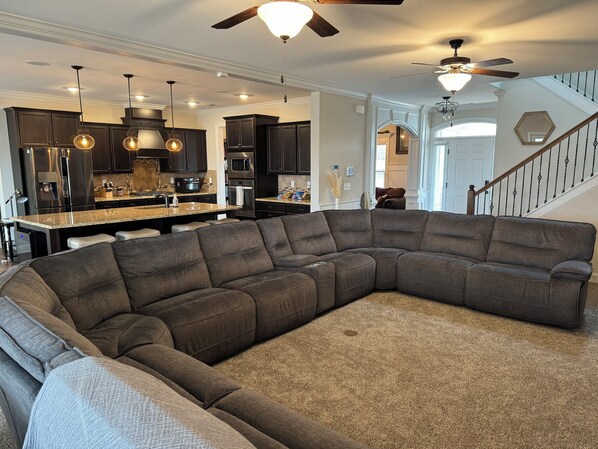 A spacious living room with a large sectional sofa, a kitchen with dark cabinets, and a staircase leading to another level. - Spacious 5-bedroom, 8-bed house in welcoming Grovetown, w/ Rogue fitness room (Grovetown)