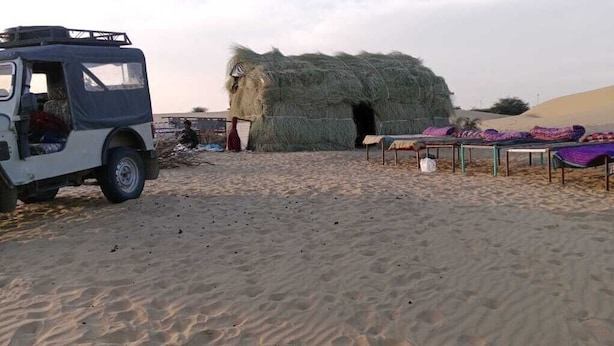 Een woestijnlandschap met een constructie van hooi, een voertuig en kleurrijke dekens op de grond.