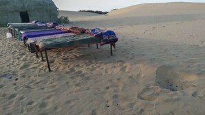 Woestijnlandschap met een rij bedden bekleed met kleurrijke dekens, een grote rotsformatie op de achtergrond en zandduinen.