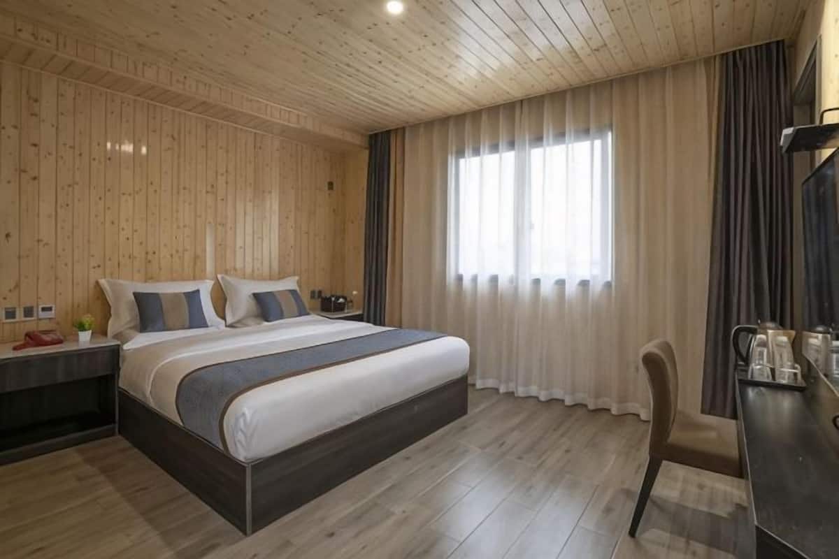 A hotel room with a wooden ceiling, a large bed with pillows, a bedside table, a chair, and a window with curtains.