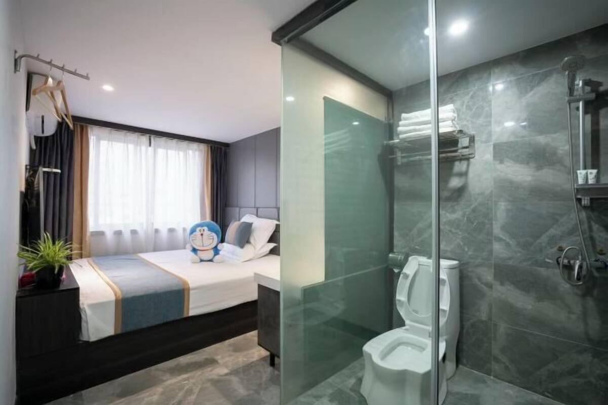 A modern hotel room with a glass-enclosed shower, a bed with a blue and white striped bedspread, and a small potted plant on the nightstand.
