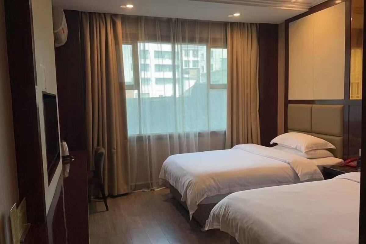 A hotel room with two beds, a large window with curtains, a wooden floor, and a wall-mounted TV.