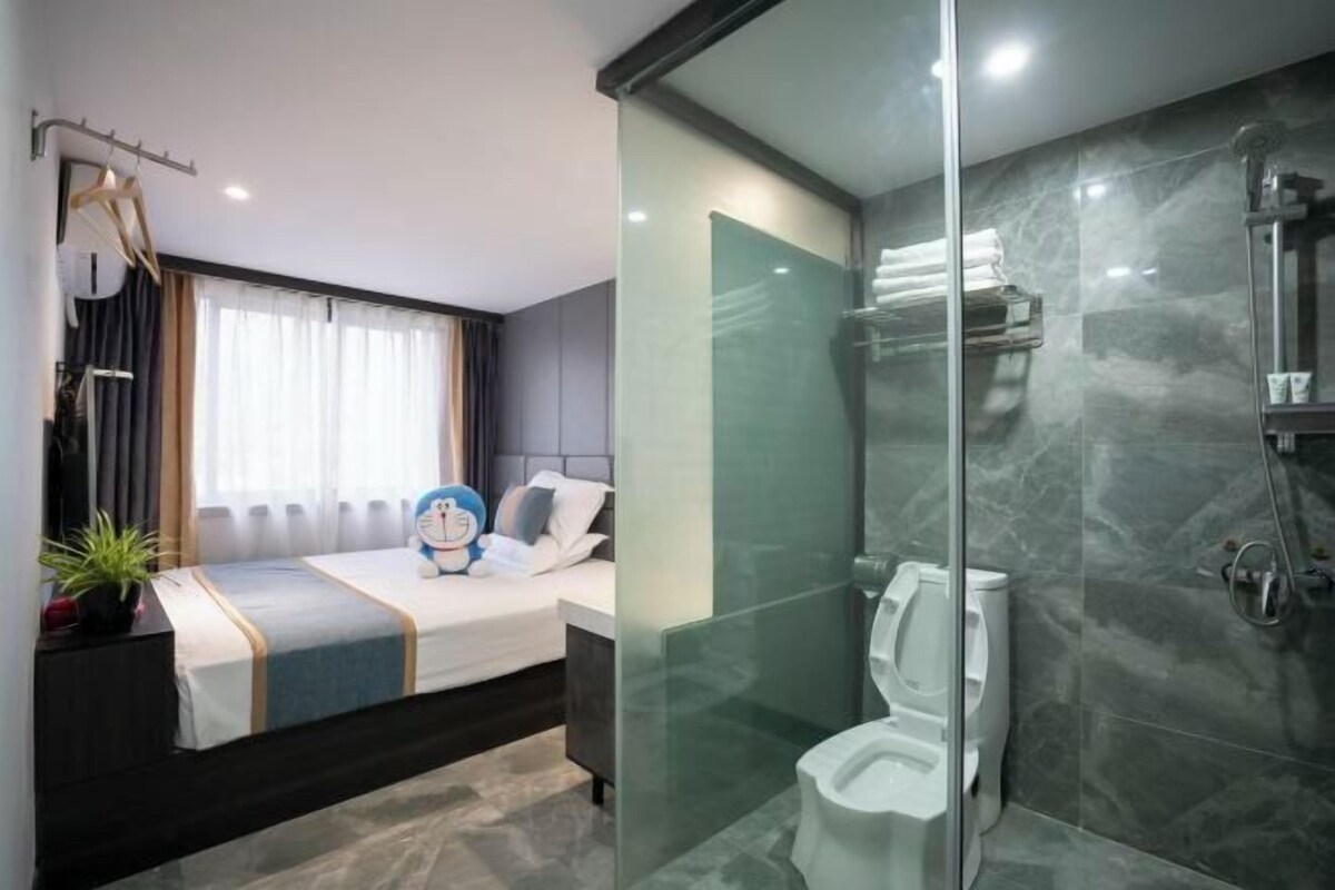 A modern hotel room with a glass-enclosed shower, a bed with a blue and white striped bedspread, and a bedside table with a plant.