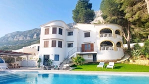 Una amplia villa blanca con piscina, balcón y montañas al fondo.