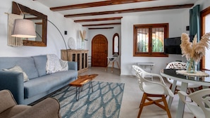 Una sala de estar con un sofá azul, una mesa de centro de madera y un tapete estampado. Se observa un comedor con sillas blancas y una mesa de vidrio. En el fondo, una puerta de madera y un televisor son visibles.