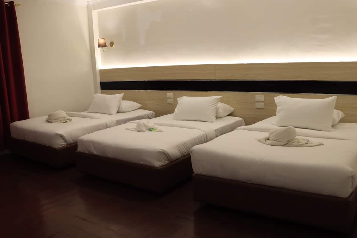 A hotel room with three single beds, each with white bedding and a towel. The room has a wooden floor and a wall-mounted light fixture.
