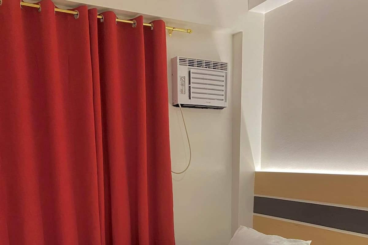 A neatly made bed with white linens and pillows, a red curtain, and an air conditioning unit on the wall.
