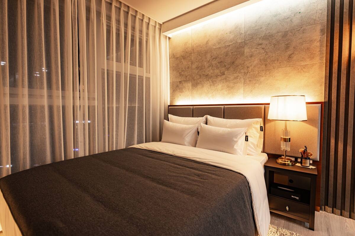 A hotel room with a large bed, a nightstand with a lamp, and a view of the city through sheer curtains.