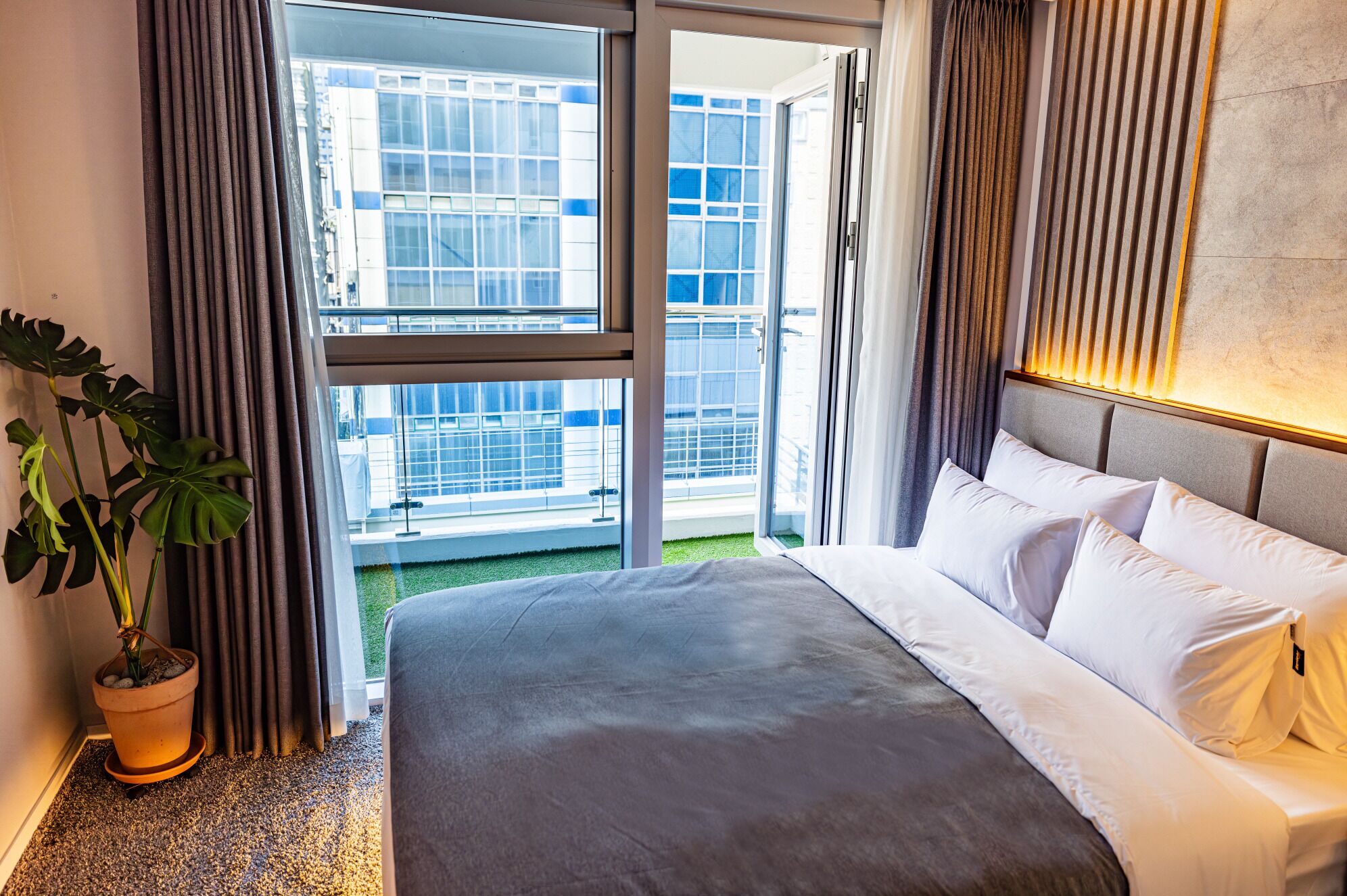 Une chambre d’hôtel avec un grand lit, une plante en pot et une vue sur un immeuble par une porte vitrée.