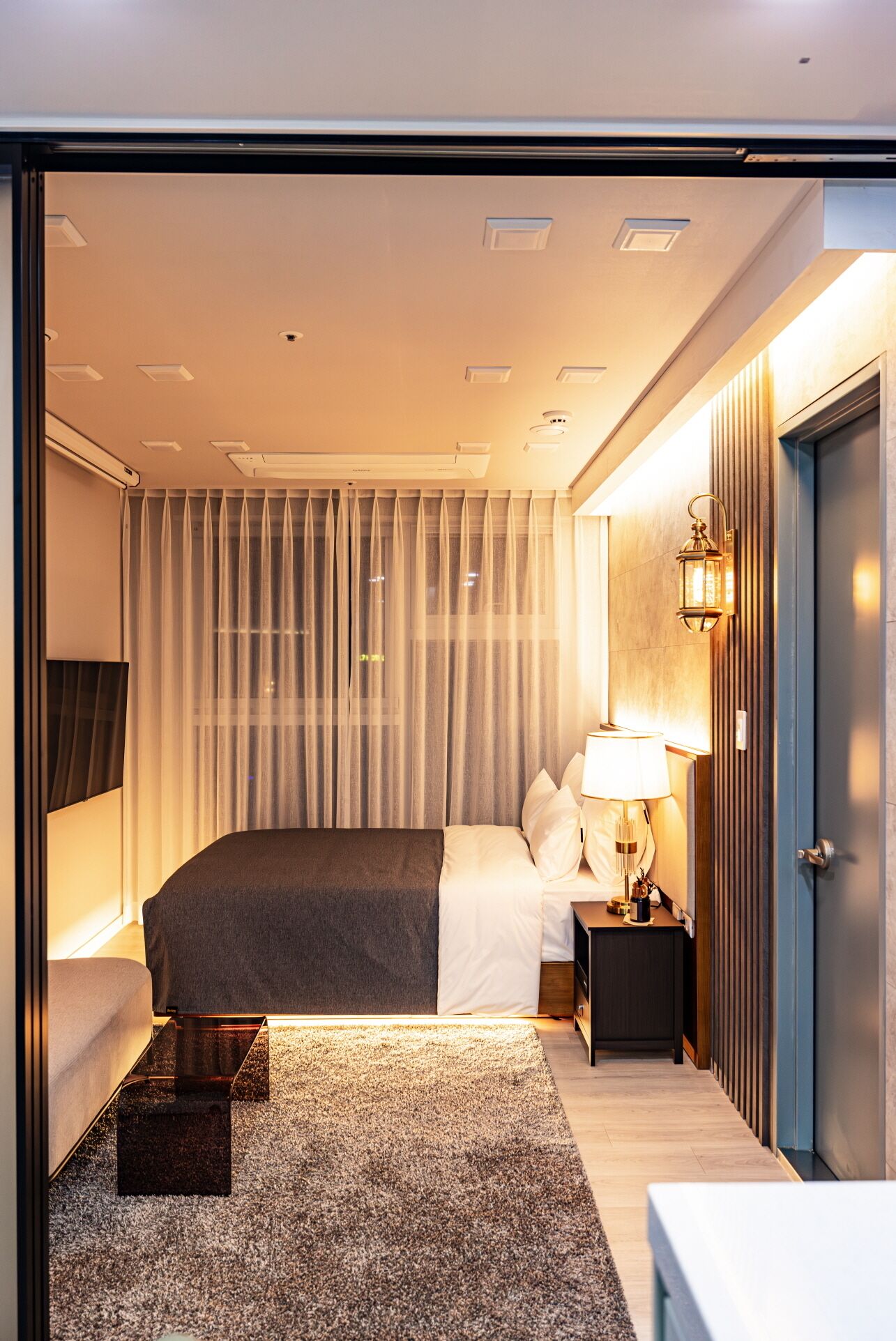 Ett modernt hotellrum med en stor säng, ett nattduksbord med en lampa, ett litet bord med en kaffekanna och en dörr med ett handtag.