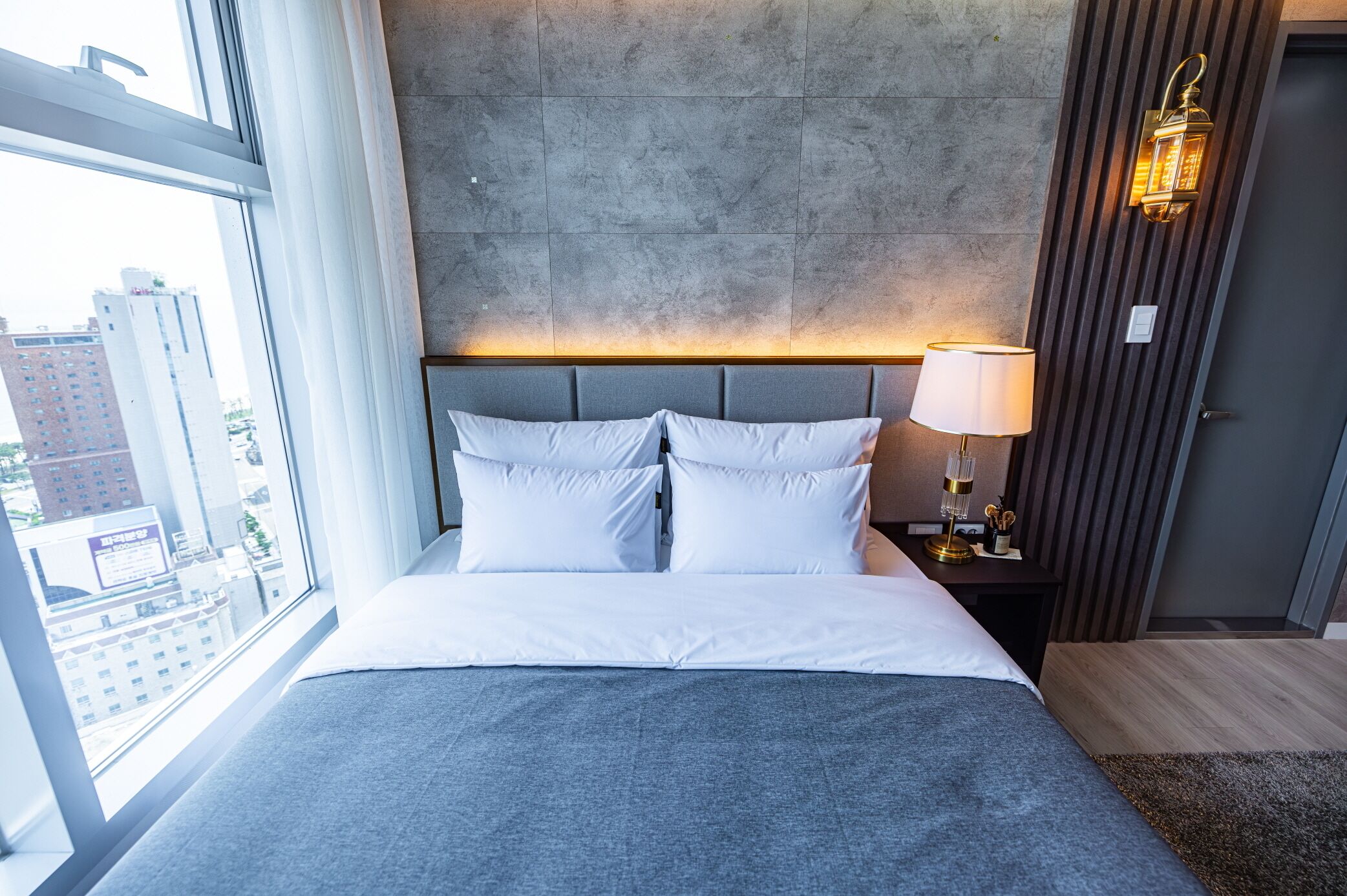 Une chambre d’hôtel moderne avec un grand lit, une table de chevet avec une lampe et une vue sur la ville grâce à une grande fenêtre.