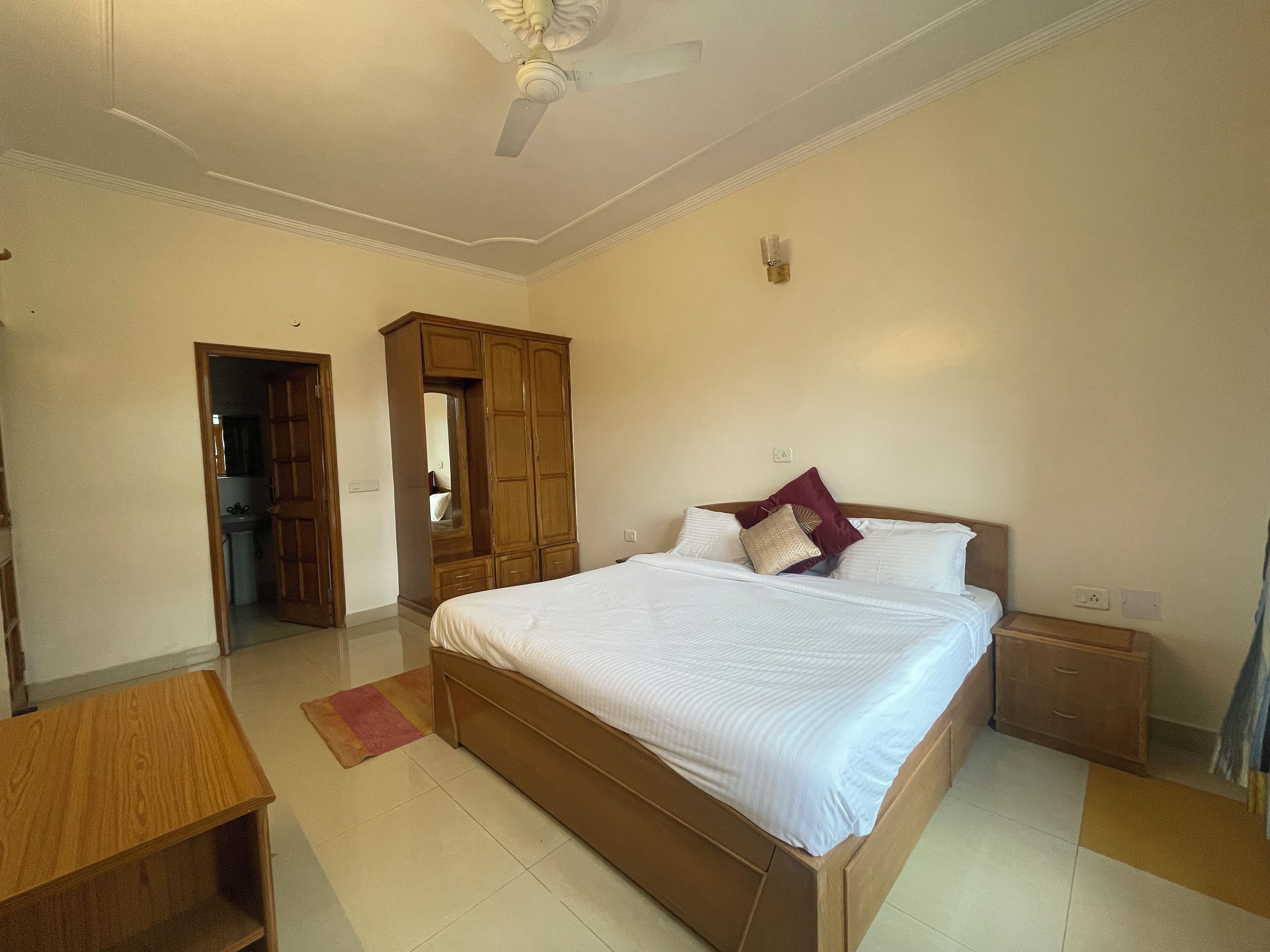 A bedroom with a bed, nightstands, a wooden wardrobe, a ceiling fan, and a door leading to another room.