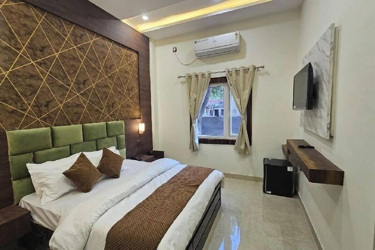 A hotel room with a bed, a TV mounted on the wall, an air conditioning unit, a window with curtains, and a small wooden shelf.