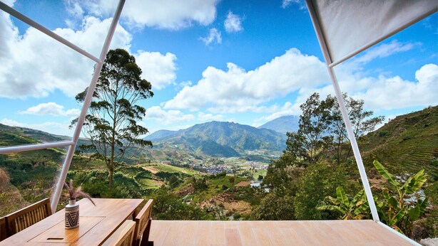 A wooden table with a vase, a bench, and a view of a valley with mountains and trees. - Luxcamp Dieng by Horison (Kejajar)