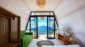 A bedroom with a bed, a green sofa, a wooden cabinet, and a large window with a mountain view. - Luxcamp Dieng by Horison (Kejajar)