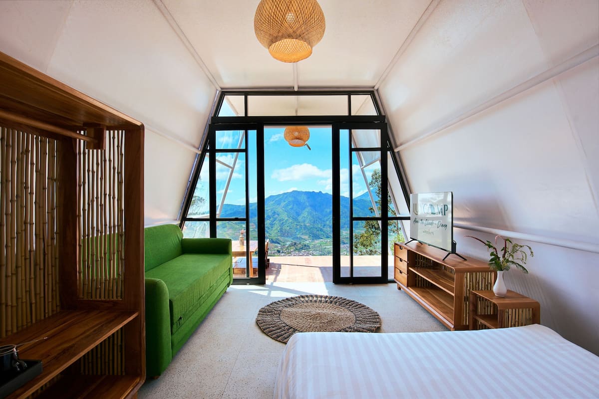 A bedroom with a bed, a green sofa, a wooden cabinet, and a large window with a mountain view.