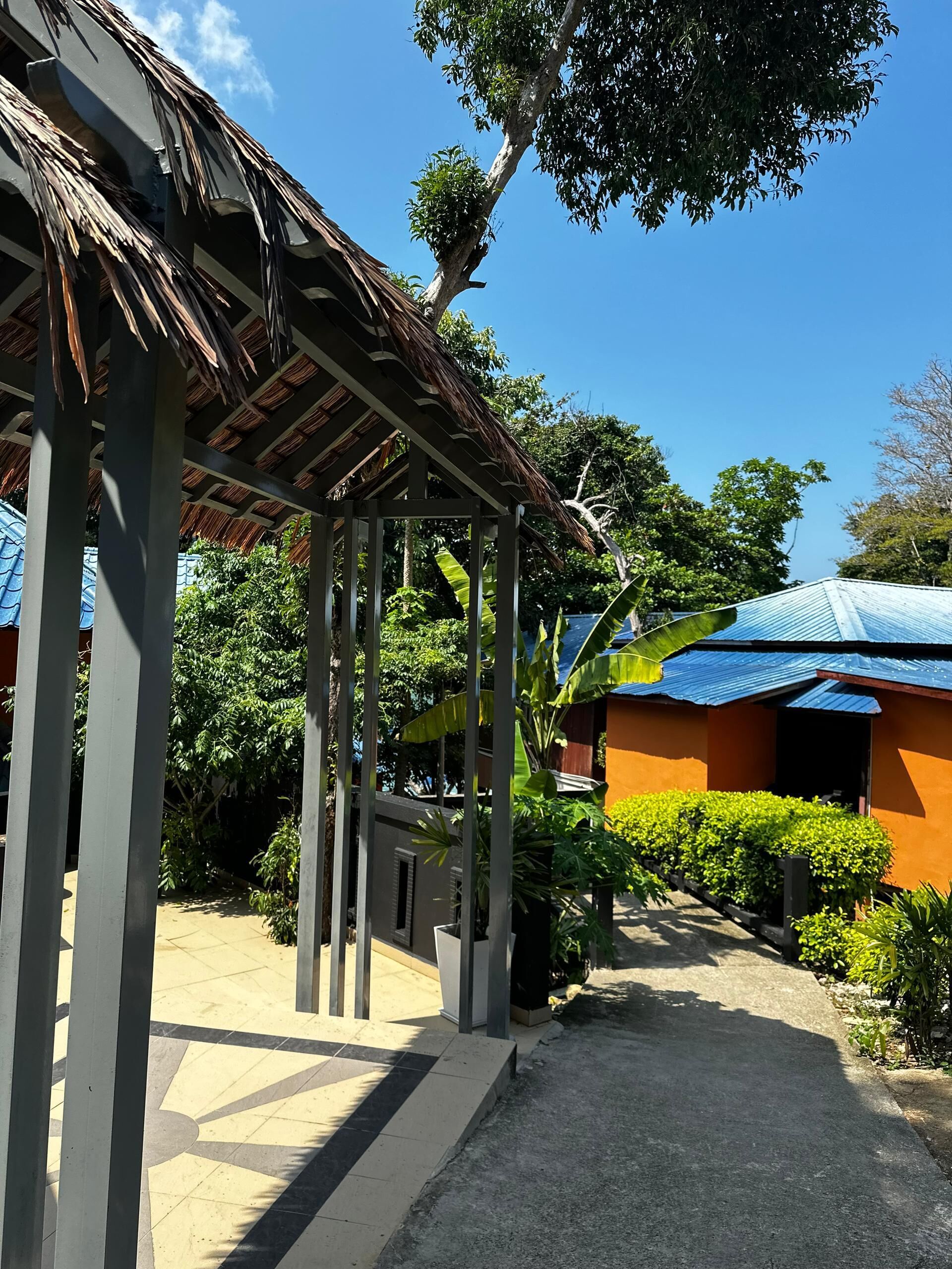 A covered outdoor area with a thatched roof structure, surrounded by lush greenery and a building with a blue roof.