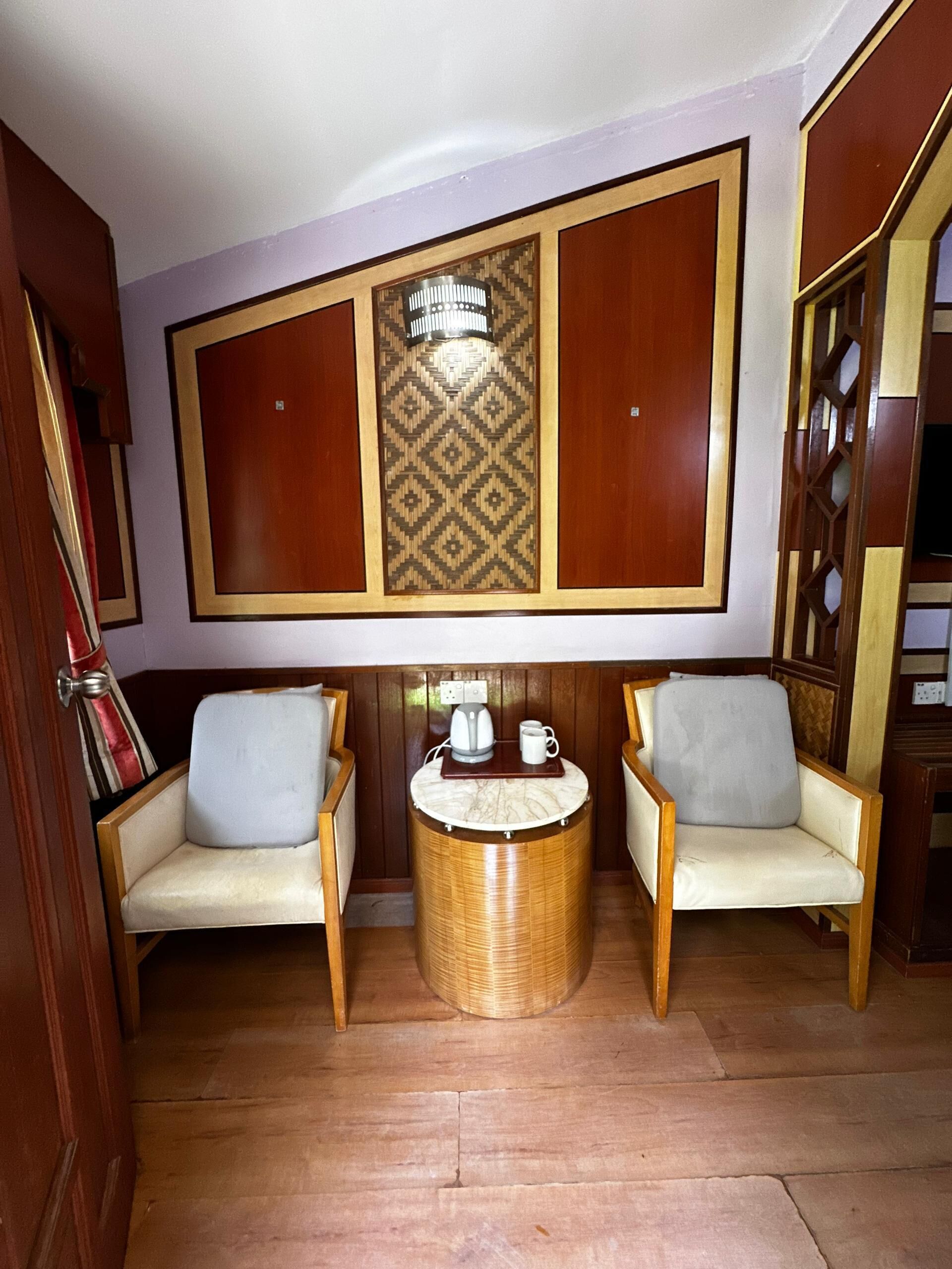 Una habitación con dos sillas de madera, una mesita redonda con una cafetera y una taza, y un panel decorativo en la pared.