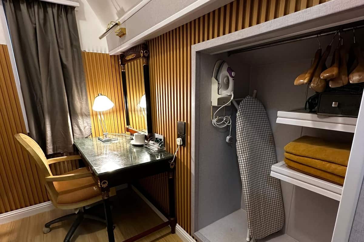 Ett kompakt hotellrum med trägolv, ett matbord med stolar, ett skrivbord med en lampa, en garderob med plats för att hänga skor och en rutig duk.