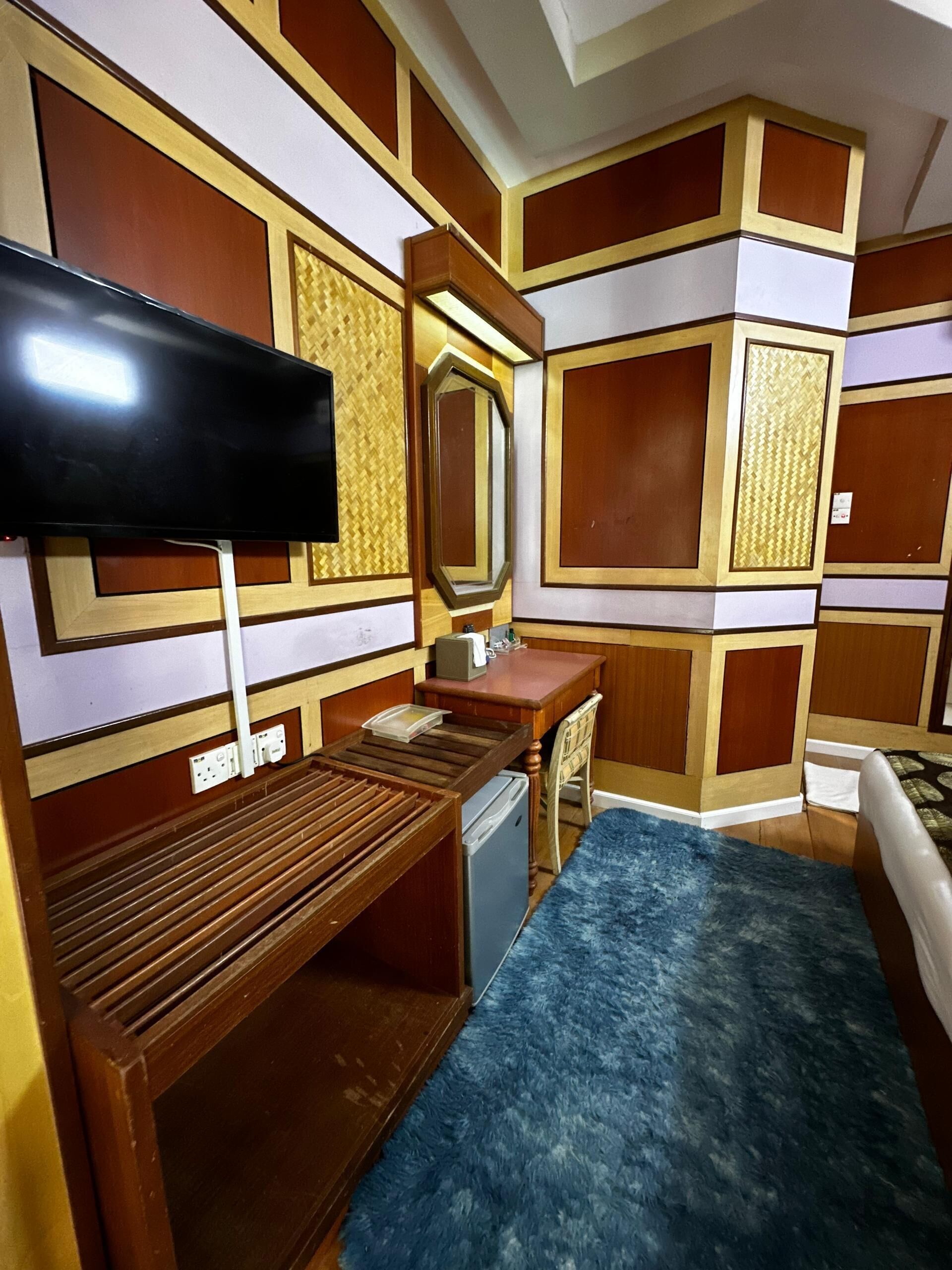 A hotel room with a wooden bench, a small desk, a television, and a large mirror.