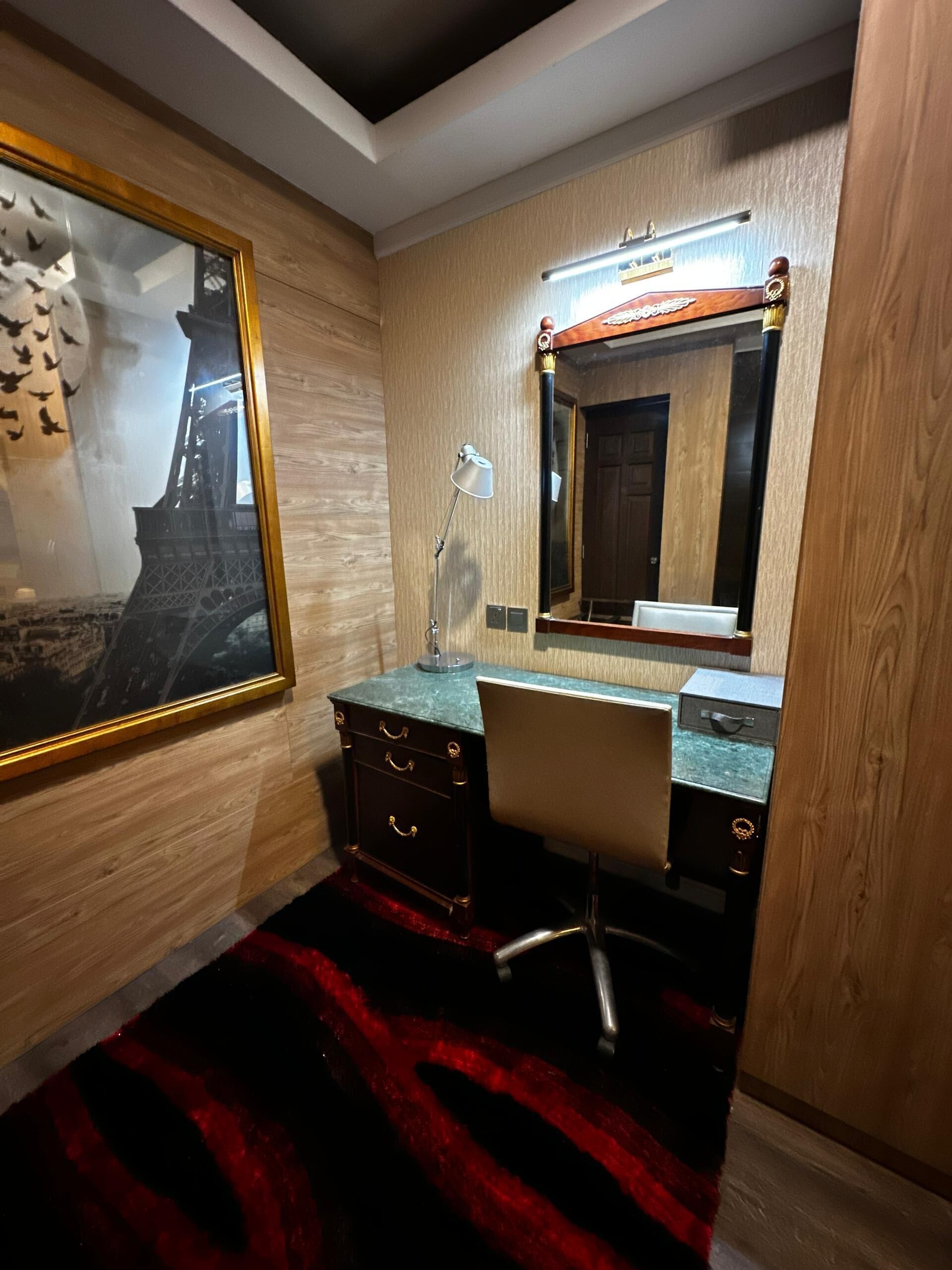 Ein Zimmer mit einer Holzwand, einem Schreibtisch mit Schubladen, einem Stuhl, einem großen Spiegel und einem roten Teppich.