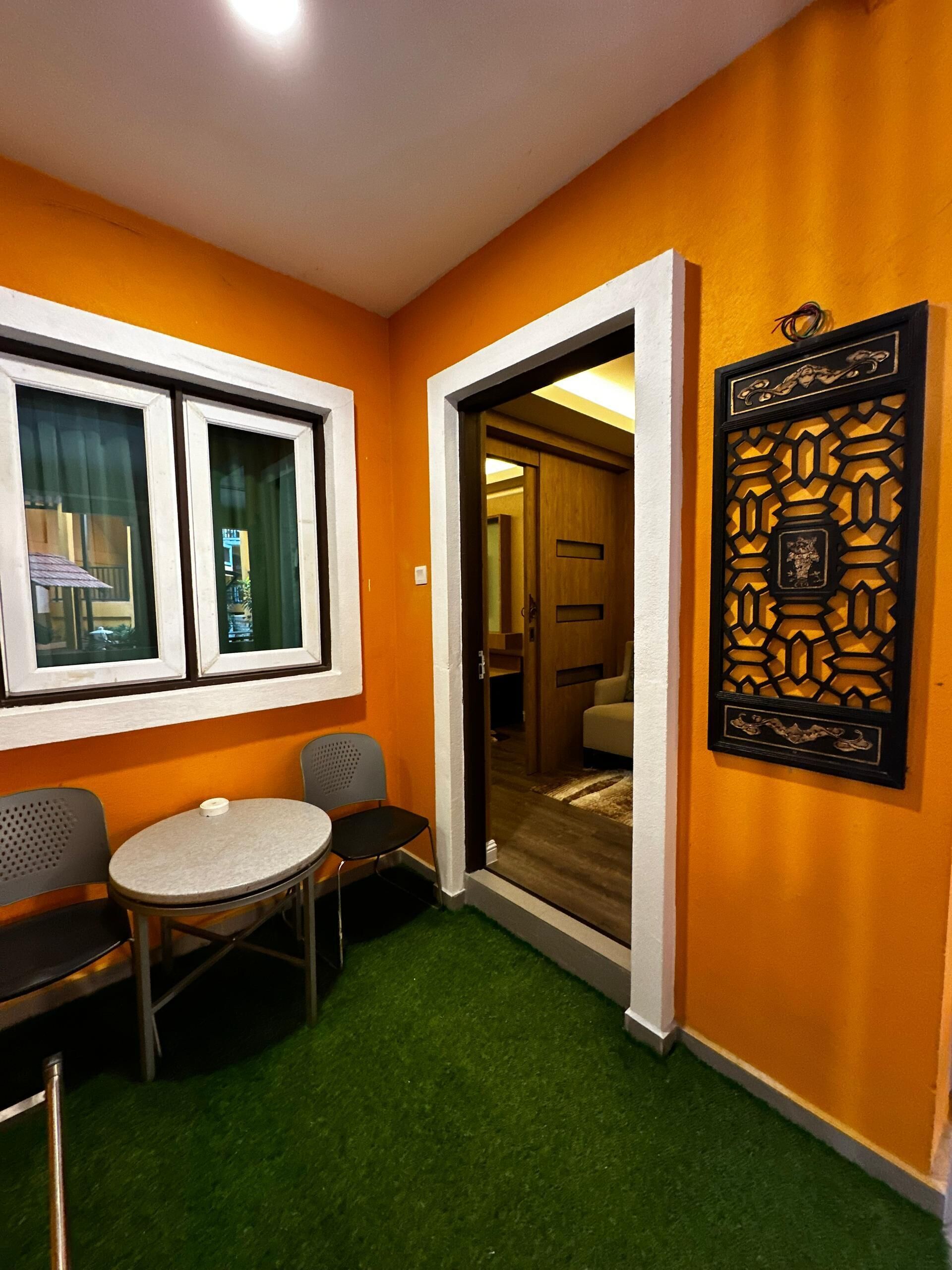Una habitación con una pared naranja, una mesa redonda pequeña con dos sillas, una ventana con cortinas y un panel decorativo.