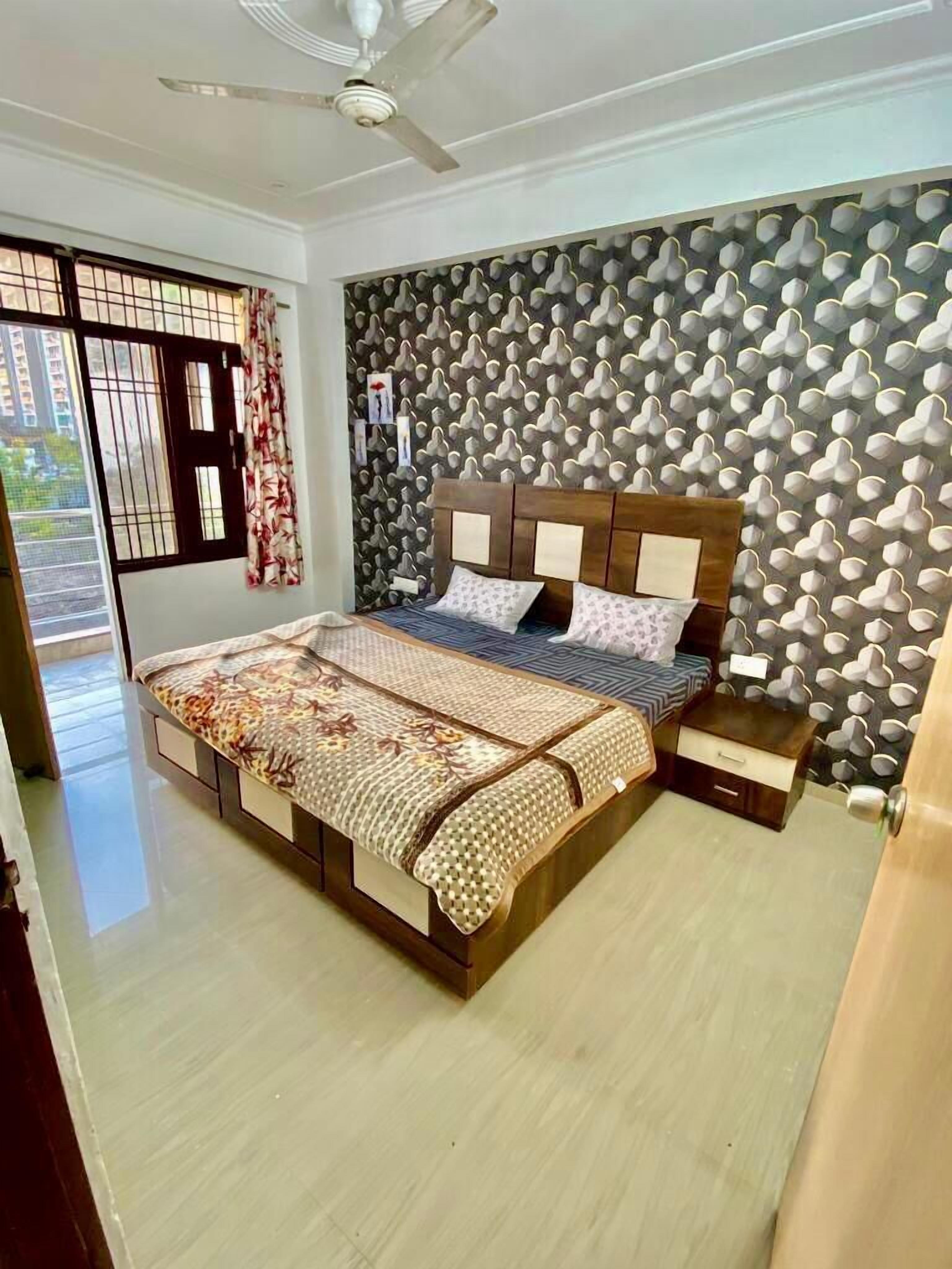 A bedroom with a bed, a nightstand, a ceiling fan, a window with curtains, and a patterned wall.