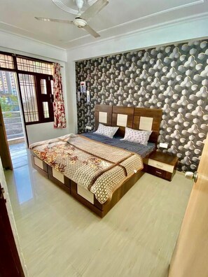 A bedroom with a bed, a nightstand, a ceiling fan, a window with curtains, and a patterned wall. - Hotel & BNB Solanki Residency Jaipur (Jaipur)