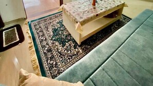 Un salón con sofá, una mesa de centro con un centro de flores y un tapete estampado. Se ve un balcón con plantas y una zona de cocina al fondo.