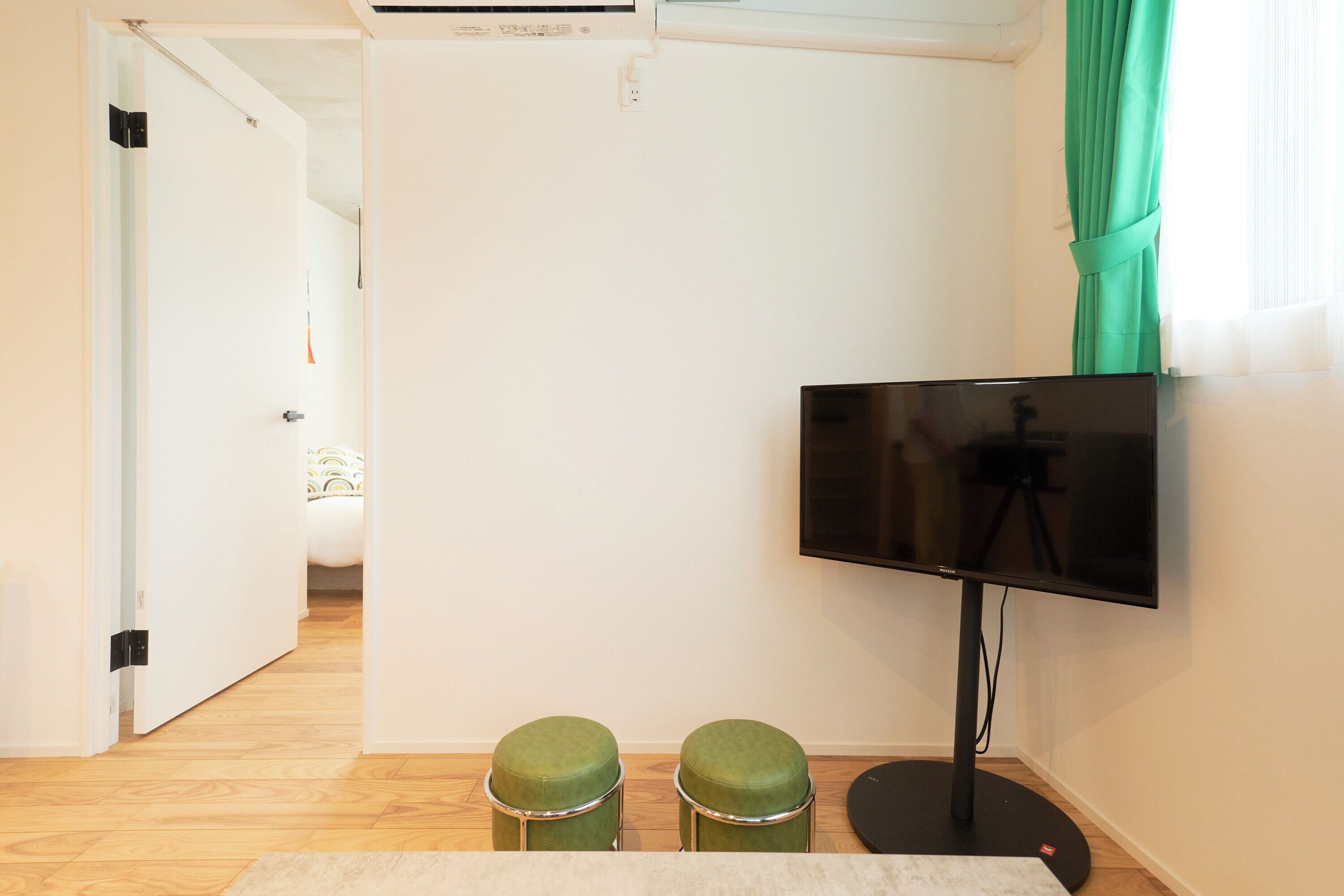 A room with a flat-screen TV on a stand, two green stools, a white door leading to another room, and a window with green curtains.