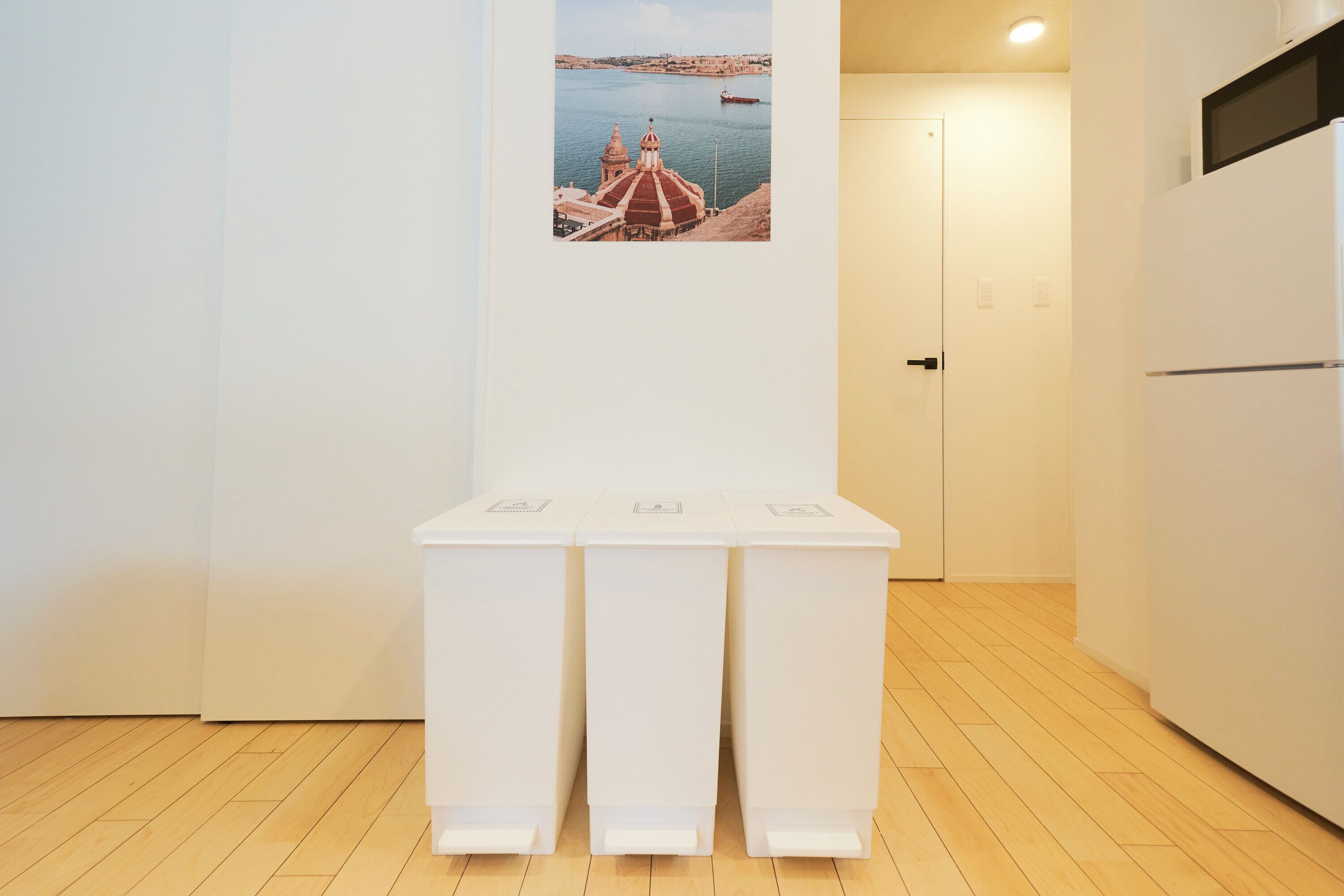 Una cocina con piso de madera, gabinetes blancos, un refrigerador y un cuadro colgado en la pared que muestra una escena costera.