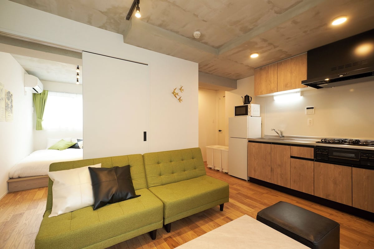 A compact living space with a green sofa, a kitchen area with wooden cabinets, a white refrigerator, and a visible bed in the background.