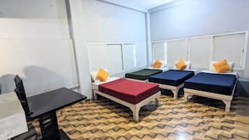 A room with four single beds, each with a different colored bedspread and matching pillows. The room has a tiled floor and white walls with a window.