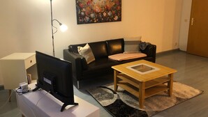 A living room with a black sofa, a wooden coffee table, a TV on a stand, a painting on the wall, and a lamp.