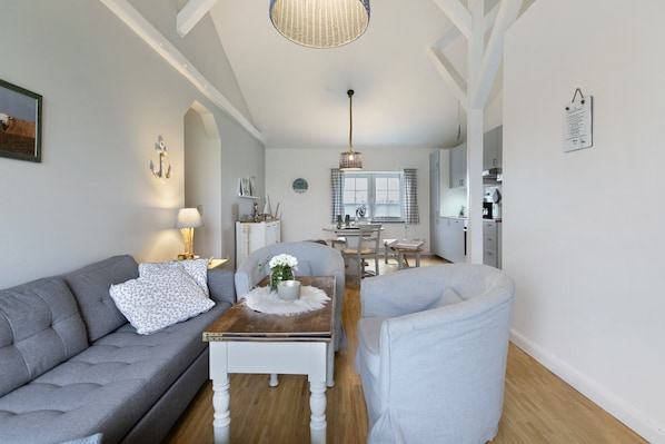 Una sala de estar moderna con un sofá, un sill ón y una mesa de centro. En el fondo, hay una zona de comedor con una mesa y sillas.